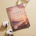 A thin printable journal titled Grief & Comfort Journal lying on a beige background with cotton flowers and a glass of water.